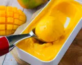 Une cuillère prélève une boule de sorbet mangue crémeux d'une boîte. Des morceaux de mangue fraîche sont visibles à côté.