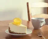 Une part de le meilleur cheesecake au citron, garnie d'une tranche de citron, sur une assiette blanche, à côté d'une tasse et d'une fourchette sur une table en bois.