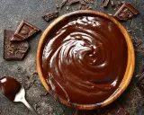 Un bol en bois déborde de chocolat fondu, prêt à faire du chocolat. Des éclats de chocolat noir et une cuillère en argent complètent la scène gourmande.