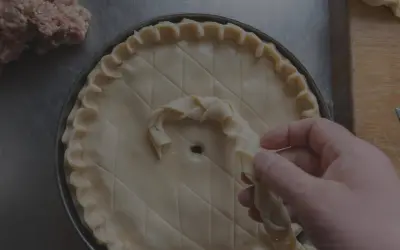 Main d'un pâtissier décorant une tarte avec une pâte brisée tressée. Un tas de farce est visible à gauche.