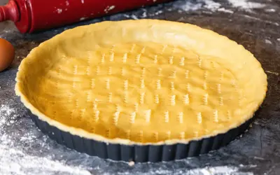 Un fond de tarte amande prêt à être garni, avec un rouleau à pâtisserie rouge et un œuf à proximité.
