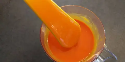 Une spatule verse un glaçage miroir orange vif dans un récipient en verre.