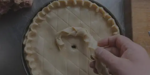 Main d'un pâtissier décorant une tarte avec une pâte brisée tressée. Un tas de farce est visible à gauche.