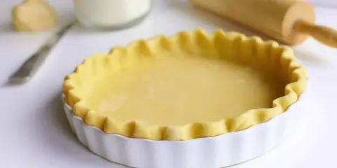 Pâte brisée prête pour la cuisson dans un plat à tarte blanc, avec un rouleau à pâtisserie en arrière-plan.