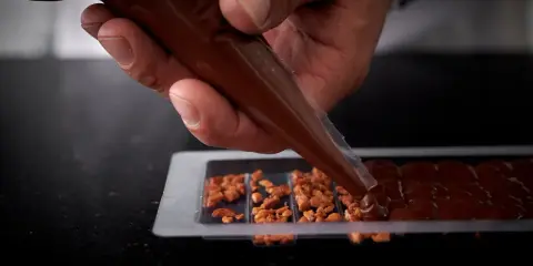 Main d'un artisan versant du chocolat fondu dans un moule pour créer une tablette de chocolat maison garnie de noisettes caramélisées.