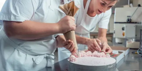 Deux pâtissières décorent un gâteau rose lors d'un cours de pâtisserie en ligne. L'une utilise une poche à douille, l'autre des fruits.