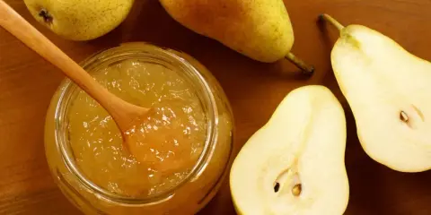 Un pot de confiture de poires maison, avec une cuillère en bois, entouré de poires fraîches coupées et entières sur fond de bois.