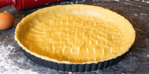 Un fond de tarte amande prêt à être garni, avec un rouleau à pâtisserie rouge et un œuf à proximité.