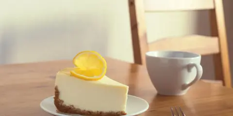 Une part de le meilleur cheesecake au citron, garnie d'une tranche de citron, sur une assiette blanche, à côté d'une tasse et d'une fourchette sur une table en bois.