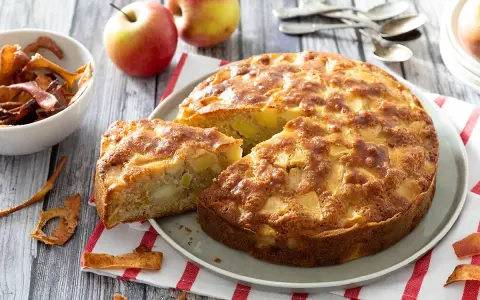 Un gâteau aux pommes super moelleux, doré et appétissant, avec des morceaux de pommes visibles. Des pommes fraîches et des chips de pommes sont disposées autour.