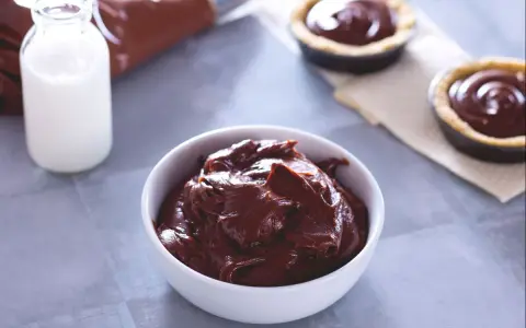 Un bol de ganache chocolat au lait onctueuse, prêt à garnir des tartelettes. Du lait et une poche à douille sont à proximité.