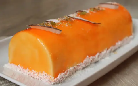 Bûche fruit exotique, un délice orangé glacé, décoré de copeaux de noix de coco et de zestes verts.