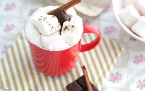 Une tasse rouge débordant de guimauves et de chocolat, parfaite pour une recette guimauve chocolat réconfortante. Un bâton de cannelle et des carrés de chocolat complètent cette gourmandise.