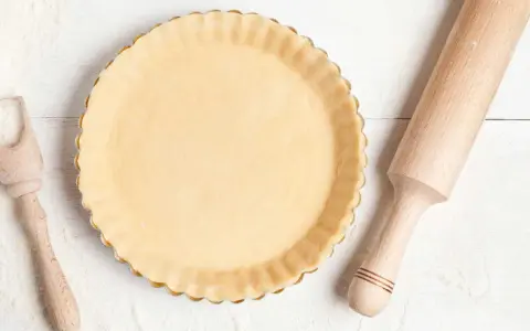 Pâte à tarte crue dans un moule, prête pour le temps de cuisson. Rouleau à pâtisserie et cuillère en bois à proximité.