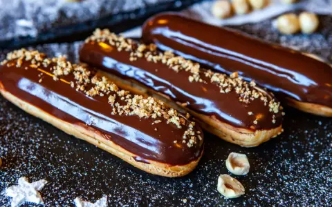 Trois éclairs au chocolat brillants, saupoudrés de noisettes concassées, sur un fond sombre parsemé de sucre glace.