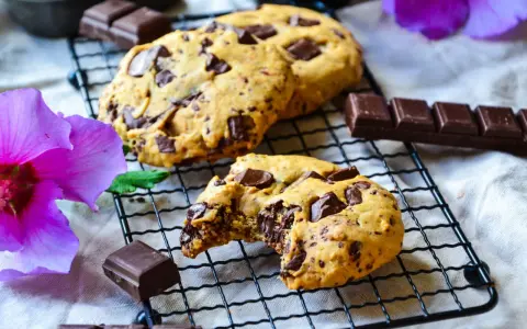 Gros plan sur des cookies chocolat faits maison, un est croqué, révélant un cœur fondant. Des morceaux de chocolat et une fleur d'hibiscus violette complètent la scène.