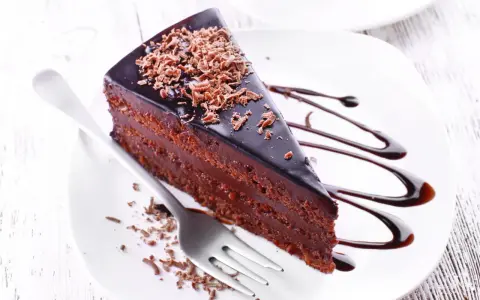 Une part de gâteau aux chocolats, nappée de glaçage brillant et garnie de copeaux de chocolat, est servie sur une assiette blanche avec un filet de sauce chocolat.