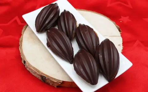 Six financiers chocolat, en forme de fèves de cacao, sur une assiette blanche, posée sur une planche de bois, sur un tissu rouge étoilé.
