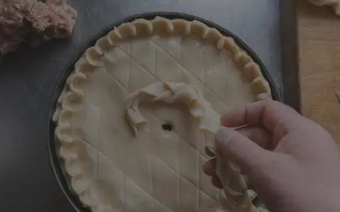 Main d'un pâtissier décorant une tarte avec une pâte brisée tressée. Un tas de farce est visible à gauche.
