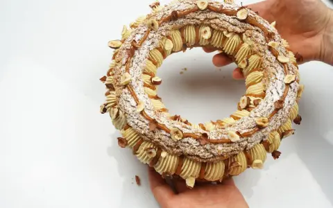 Un Paris-Brest craquelin, orné de noisettes et de caramel, est tenu par des mains.
