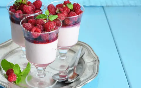 Trois panna cotta onctueuses, garnies de framboises fraîches et d'un coulis de framboise, sur un plateau argenté.
