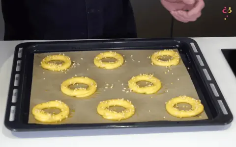Huit ronds de pâte à choux, prêts à devenir un délicieux Paris-Brest, sont saupoudrés d'amandes effilées sur une plaque de cuisson.