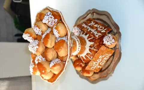Une pièce montée de choux garnis de crème, nappés de caramel et décorés de perles blanches. Sur un plateau, d'autres choux et des tuiles aux amandes portent des inscriptions.