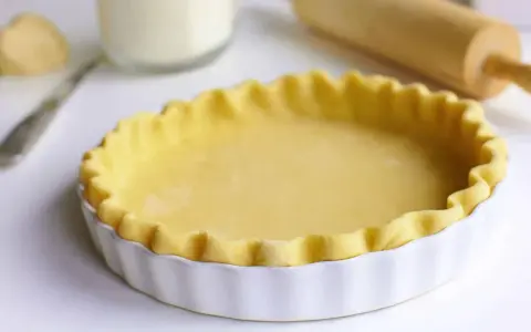 Pâte brisée prête pour la cuisson dans un plat à tarte blanc, avec un rouleau à pâtisserie en arrière-plan.
