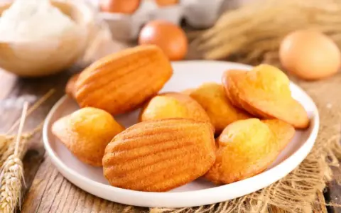 Des madeleines dorées, parfaites pour une recette gâteau madeleine meilleur pâtissier. Des œufs et de la farine en arrière-plan.