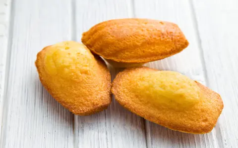 Trois madeleines dorées, tout juste sorties du four, sur une planche en bois. La cuisson madeleine est réussie, leur texture est parfaite.