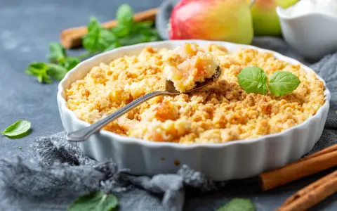 Un délicieux crumble aux pommes à l'ancienne, garni de feuilles de menthe fraîche, avec des pommes et des bâtons de cannelle en arrière-plan.