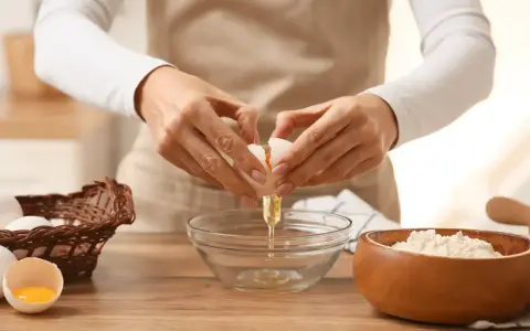 Préparation d'une recette pâte brisée facile : une main casse un œuf dans un bol, à côté d'un panier d'œufs et d'un bol de farine.