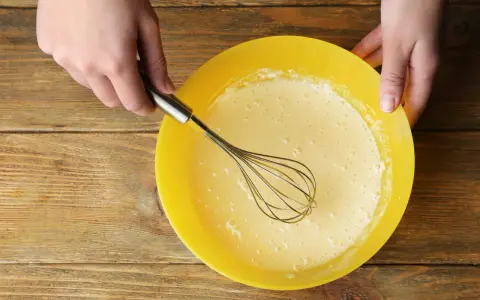 Mains préparant la pâte à crêpes dans un bol jaune. La pâte est prête pour son temps de repos.
