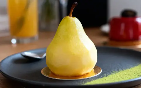 Un entremet poire, glacé et brillant, repose sur une assiette noire avec une touche de poudre verte.