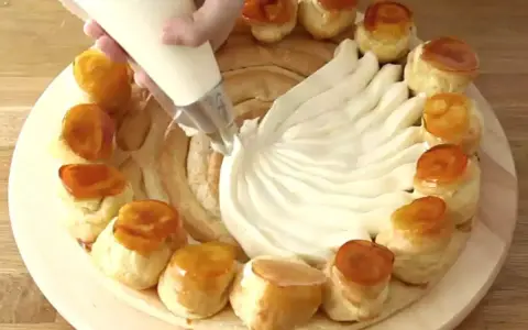 Une main garnit une couronne de choux avec de la crème chiboust, créant un dessert spectaculaire.