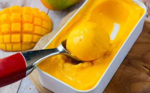 Une cuillère prélève une boule de sorbet mangue crémeux d'une boîte. Des morceaux de mangue fraîche sont visibles à côté.
