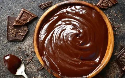 Un bol en bois déborde de chocolat fondu, prêt à faire du chocolat. Des éclats de chocolat noir et une cuillère en argent complètent la scène gourmande.