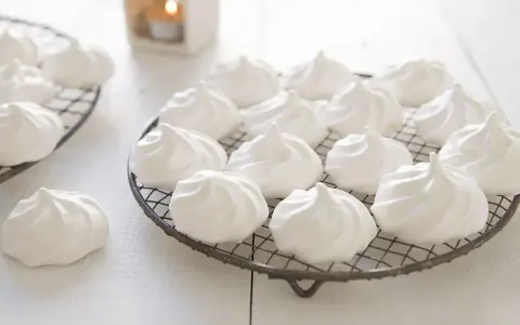 Douces meringues blanches, prêtes pour la cuisson. Elles reposent sur des grilles, évoquant la douceur et la légèreté.