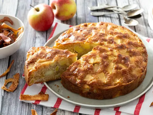 Un gâteau aux pommes super moelleux, doré et appétissant, avec des morceaux de pommes visibles. Des pommes fraîches et des chips de pommes sont disposées autour.