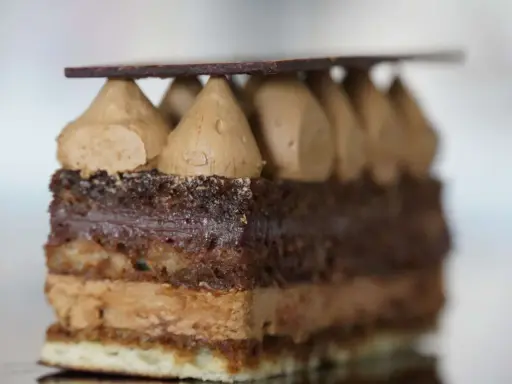 Une tranche de gâteau Opéra, avec sa composition de couches de biscuit Joconde, ganache au chocolat et crème au café, surmontée de dômes de crème et d'une fine plaque de chocolat.