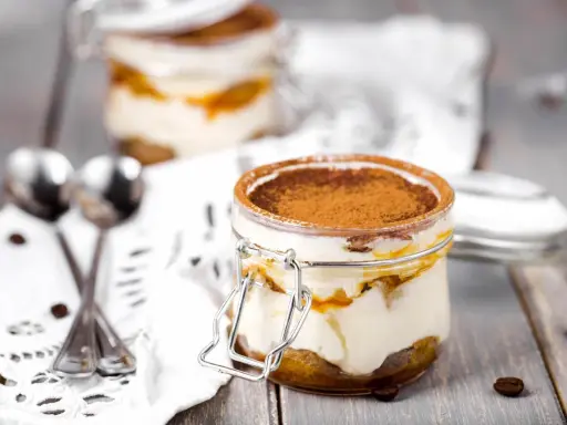 Deux bocaux de tiramisu, le meilleur dessert, saupoudrés de cacao, avec des grains de café et des cuillères sur une nappe en dentelle.