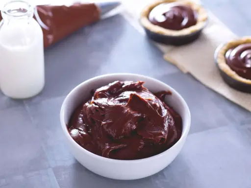 Un bol de ganache chocolat au lait onctueuse, prêt à garnir des tartelettes. Du lait et une poche à douille sont à proximité.