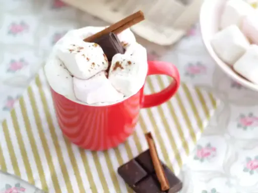 Une tasse rouge débordant de guimauves et de chocolat, parfaite pour une recette guimauve chocolat réconfortante. Un bâton de cannelle et des carrés de chocolat complètent cette gourmandise.