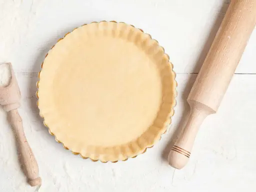 Pâte à tarte crue dans un moule, prête pour le temps de cuisson. Rouleau à pâtisserie et cuillère en bois à proximité.