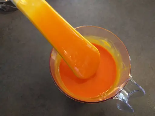 Une spatule verse un glaçage miroir orange vif dans un récipient en verre.