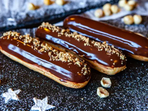 Trois éclairs au chocolat brillants, saupoudrés de noisettes concassées, sur un fond sombre parsemé de sucre glace.