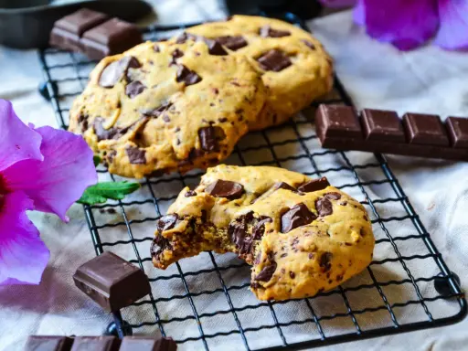 Gros plan sur des cookies chocolat faits maison, un est croqué, révélant un cœur fondant. Des morceaux de chocolat et une fleur d'hibiscus violette complètent la scène.
