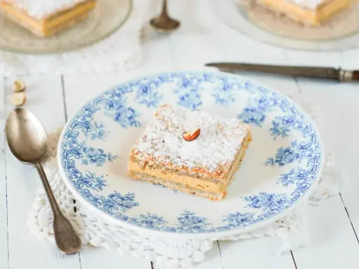 Un carré de gâteau russe, saupoudré de sucre glace et garni d'une noisette, sur une assiette ornée de bleu.