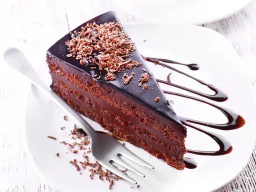Une part de gâteau aux chocolats, nappée de glaçage brillant et garnie de copeaux de chocolat, est servie sur une assiette blanche avec un filet de sauce chocolat.