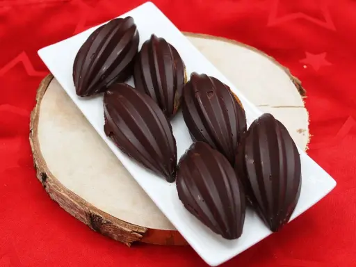 Six financiers chocolat, en forme de fèves de cacao, sur une assiette blanche, posée sur une planche de bois, sur un tissu rouge étoilé.